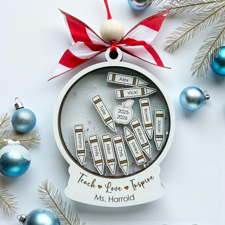 Close-up of custom engraved teacher Christmas ornament with student name crayons, silver and blue holiday background, and red ribbon bow. Shaker ornament gift idea for teachers, personalized classroom Christmas keepsake.