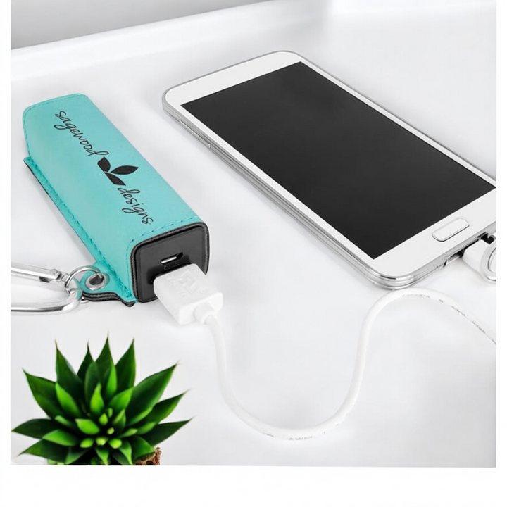 Turquoise laser-engraved leatherette 2200mAh power bank connected to a smartphone, featuring a silver carabiner and USB charging cord.